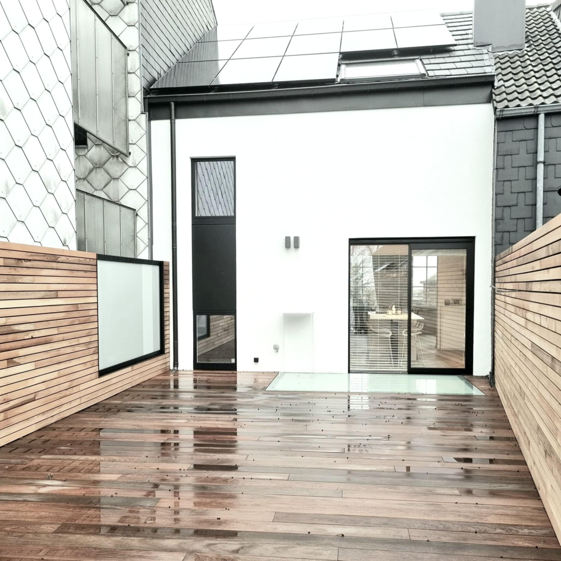 Une maison blanche moderne avec une terrasse et une clôture en bois. Des portes à cadre noir et une petite fenêtre.