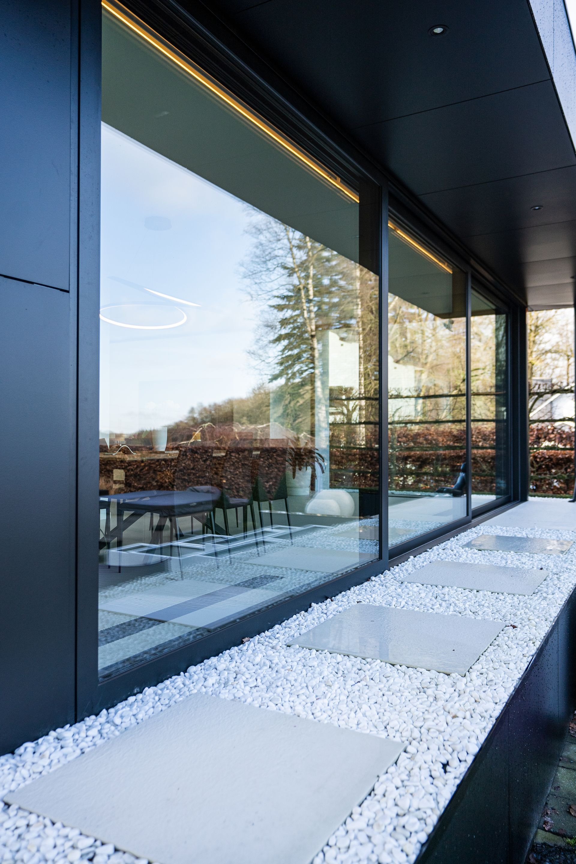 De grandes fenêtres à cadre noir reflètent une scène extérieure ensoleillée, bordée d'un lit de galets blancs et de carrés de béton.
