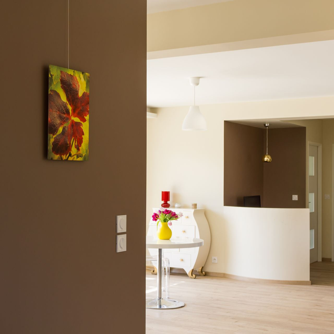 Un tableau aux couleurs vives représentant une feuille est accroché à un mur brun, donnant sur une salle à manger lumineuse.