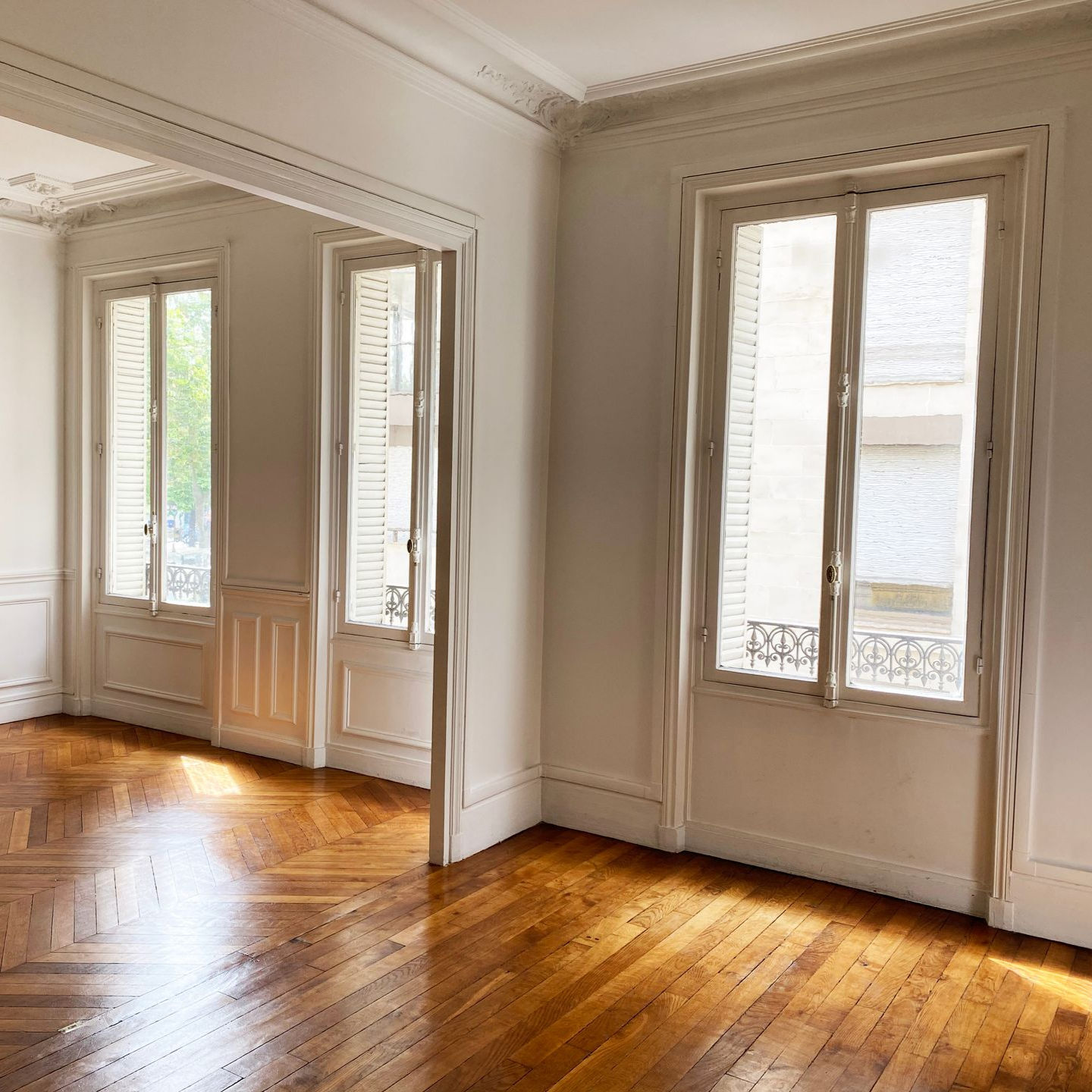 Une pièce vide avec un parquet en chevrons ciré, des murs blancs, des moulures ornementées et de hautes fenêtres.