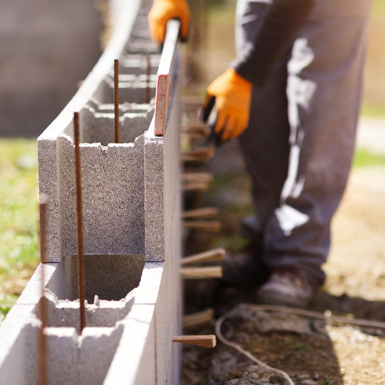 Un ouvrier du bâtiment portant des gants jaunes aligne un bloc de béton sur un mur en parpaing.