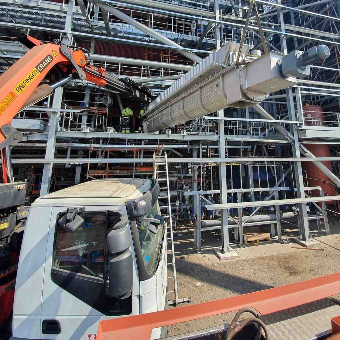 Obra en construcción con una excavadora naranja levantando una viga de acero junto a un camión blanco y andamios metálicos.