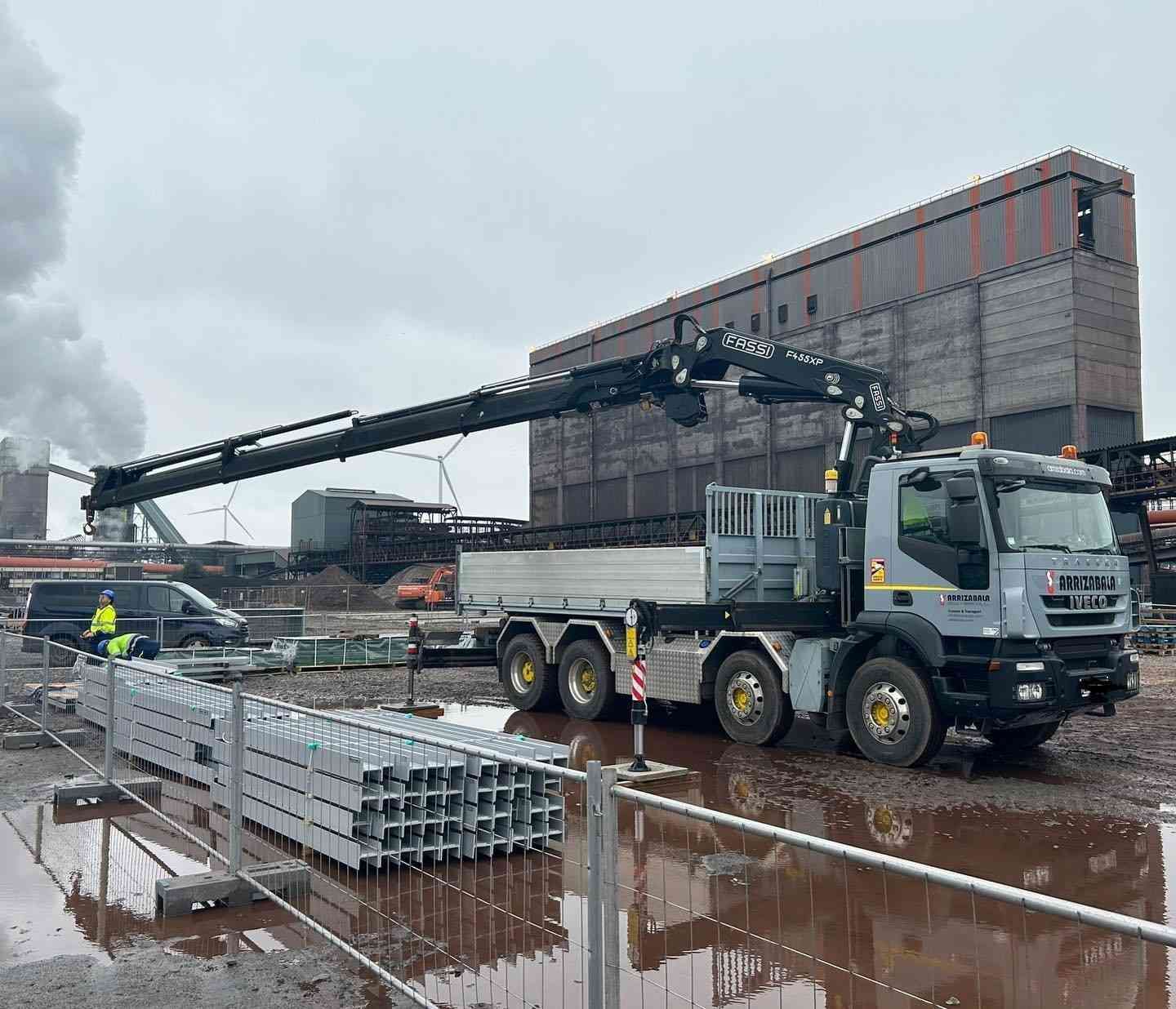 Camión grúa descargando materiales en una obra en construcción húmeda junto a un gran edificio.
