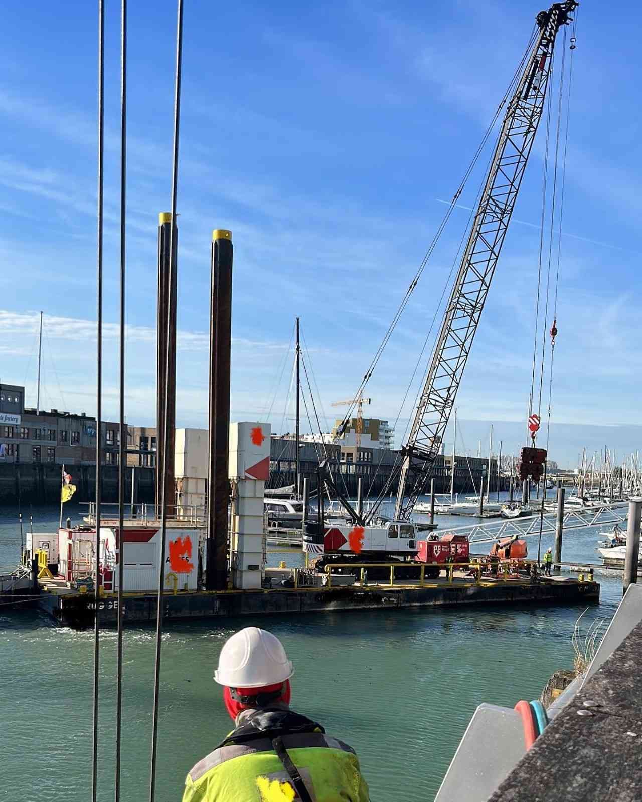 Barcaza de construcción portuaria con grúa y pilotes altos, vista desde arriba a un trabajador con casco blanco junto al agua.