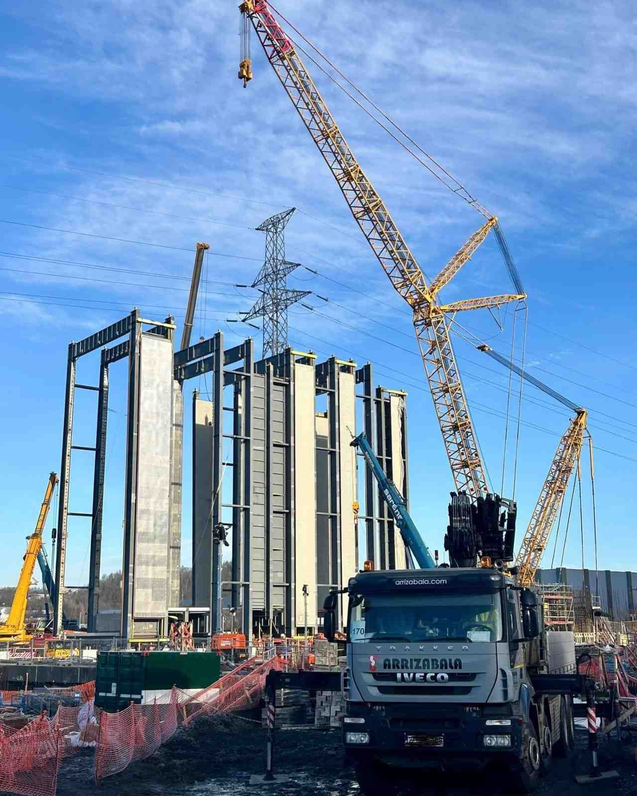 Obra en construcción con una grúa y un camión junto a una estructura de hormigón parcialmente construida bajo un cielo azul.