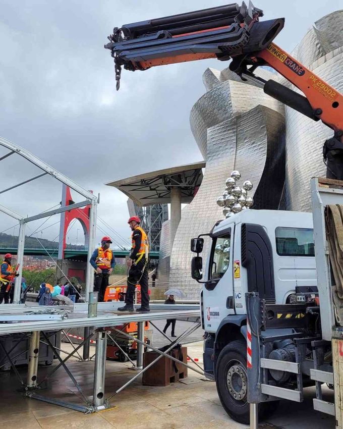 Trabajadores en un escenario junto a un camión blanco y una grúa, bajo un cielo nublado, con una escultura moderna al fondo.