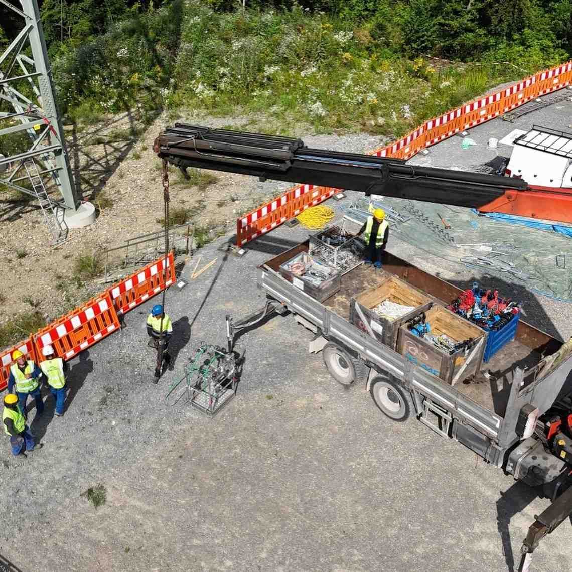 Un equipo de construcción levanta una viga negra sobre una carretera junto a un camión y barreras naranjas.