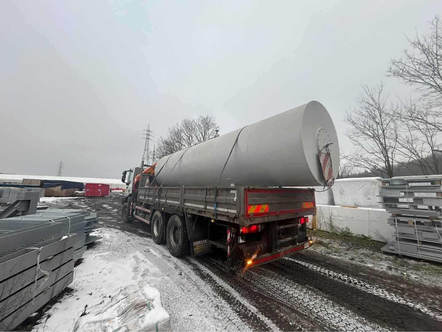 Un gran tanque plateado sobre un remolque que circula por un patio industrial nevado.
