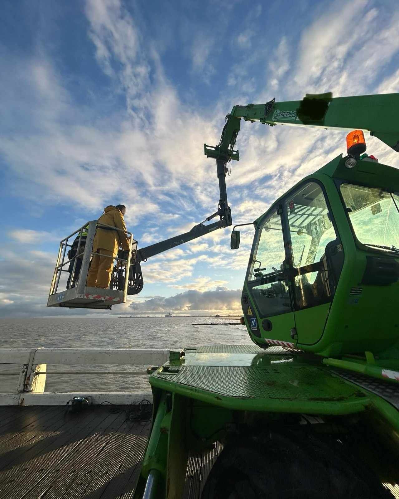 Una grúa verde levanta una máquina amarilla sobre agua helada bajo un cielo nublado.
