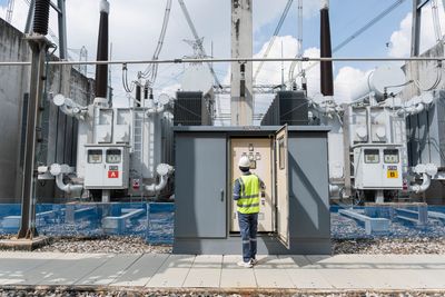 Ein Arbeiter in einer neongelben Weste steht am Eingang eines Umspannwerks, zu beiden Seiten befinden sich Transformatoren.