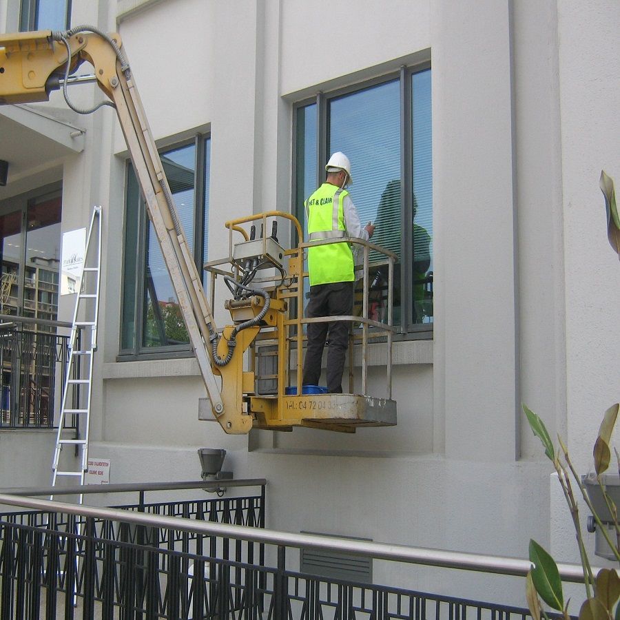 Ouvrier en gilet jaune sur une nacelle de nettoyage de vitres près d'une façade d'immeuble.
