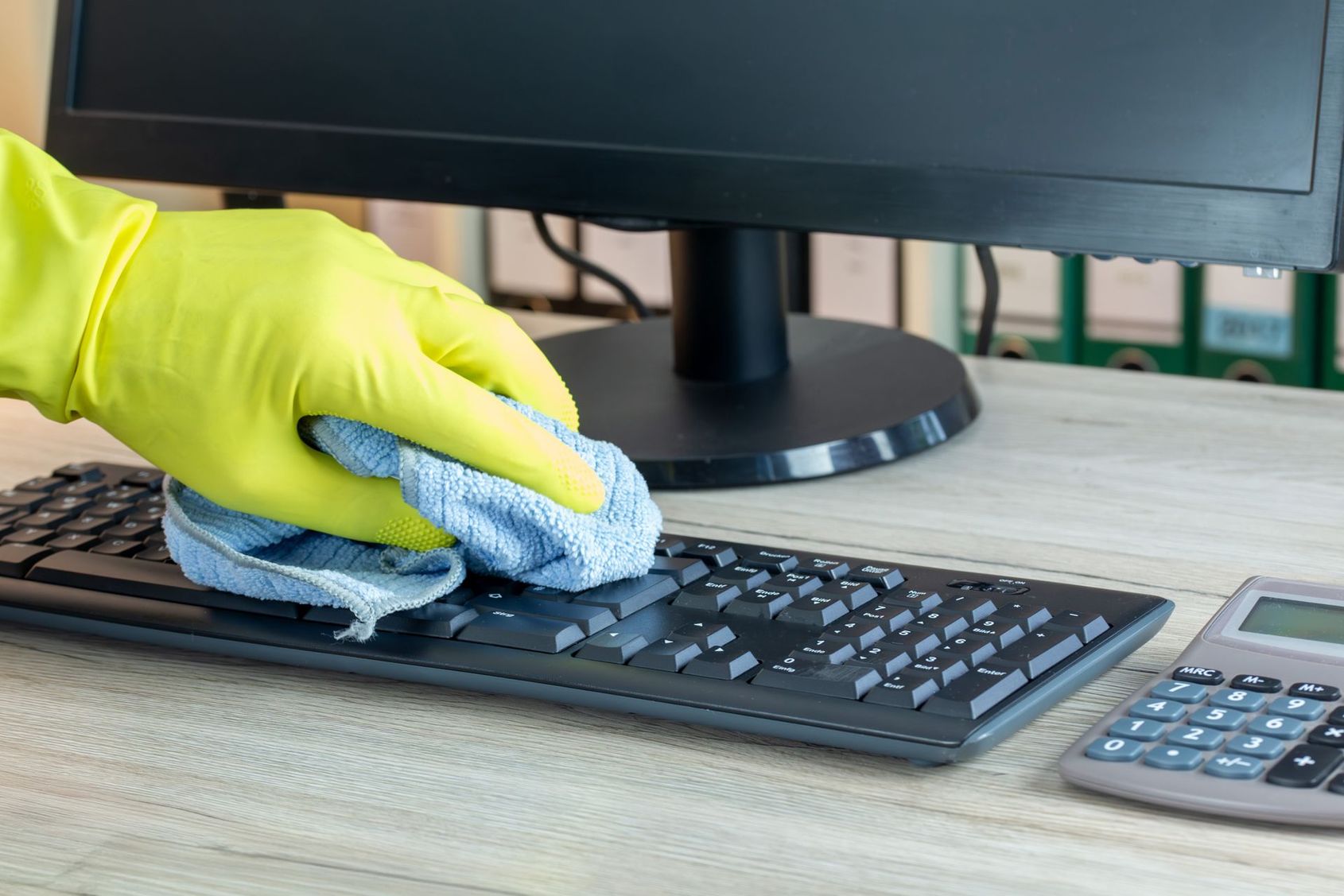Une main gantée essuie un clavier d'ordinateur avec un chiffon, à côté d'un écran d'ordinateur et d'une calculatrice.