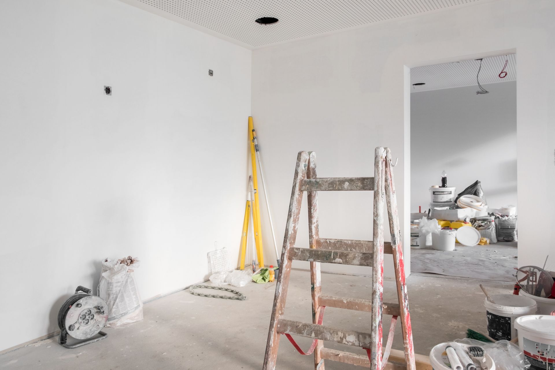 Pièce en rénovation avec échelle, outils et fournitures sur un sol blanc à côté d'une grande ouverture de miroir.