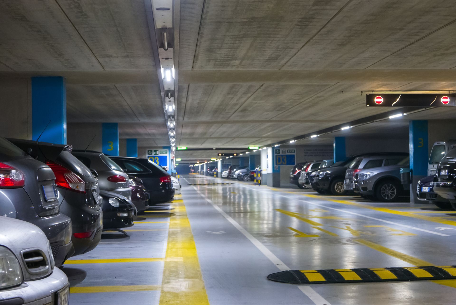 Stationnement souterrain avec des rangées de voitures garées et une voie mouillée et réfléchissante sous des néons.