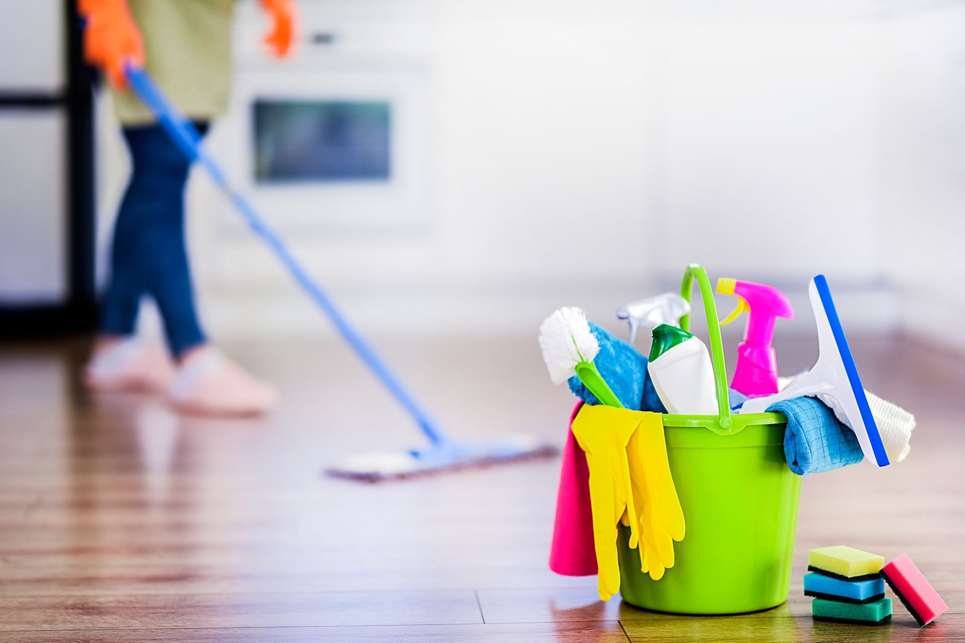 Des produits de nettoyage dans un seau vert sur un sol en bois, avec une personne qui passe la serpillière en arrière-plan.