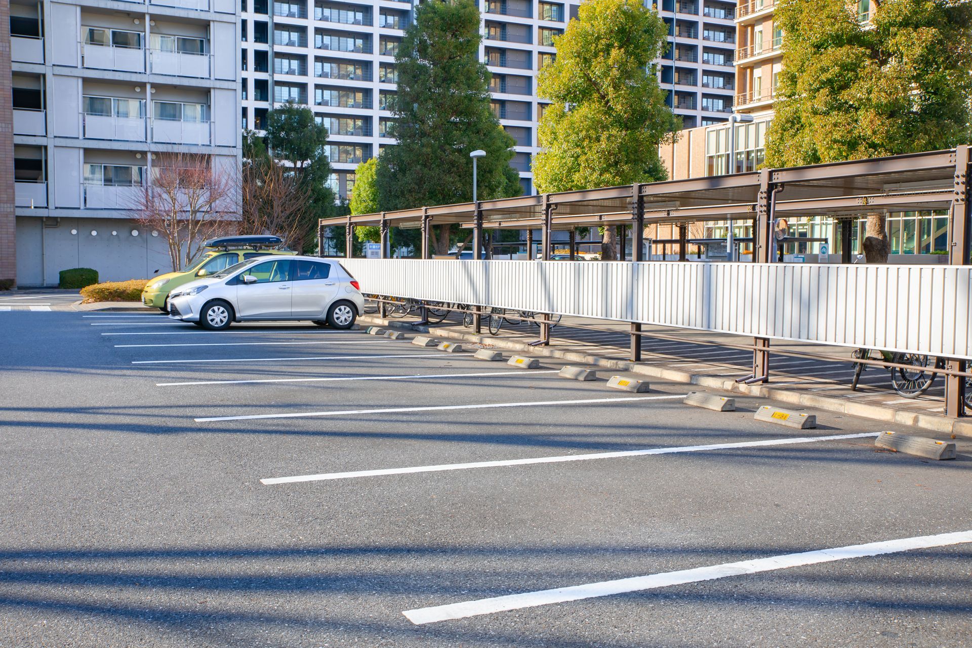 Stationnement vide à côté d'immeubles, avec une petite voiture argentée et une allée clôturée.
