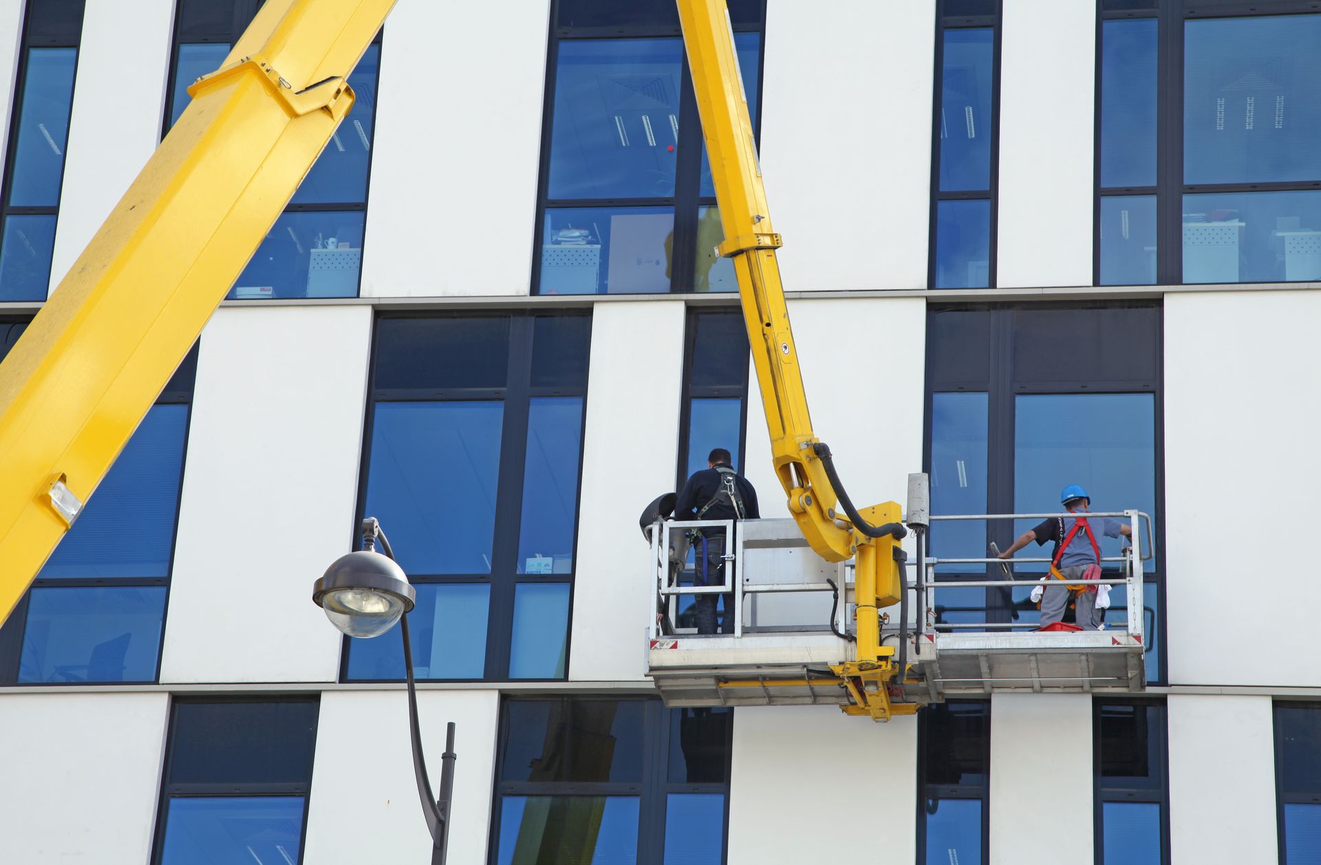 Des ouvriers, installés sur une nacelle jaune, réparent la façade d'un immeuble moderne aux fenêtres bleues.