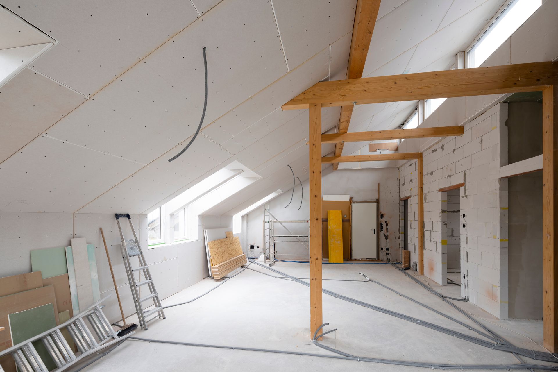 Intérieur de grenier non aménagé avec murs blancs en pente, puits de lumière, poutres en bois et échelles appuyées contre le mur.