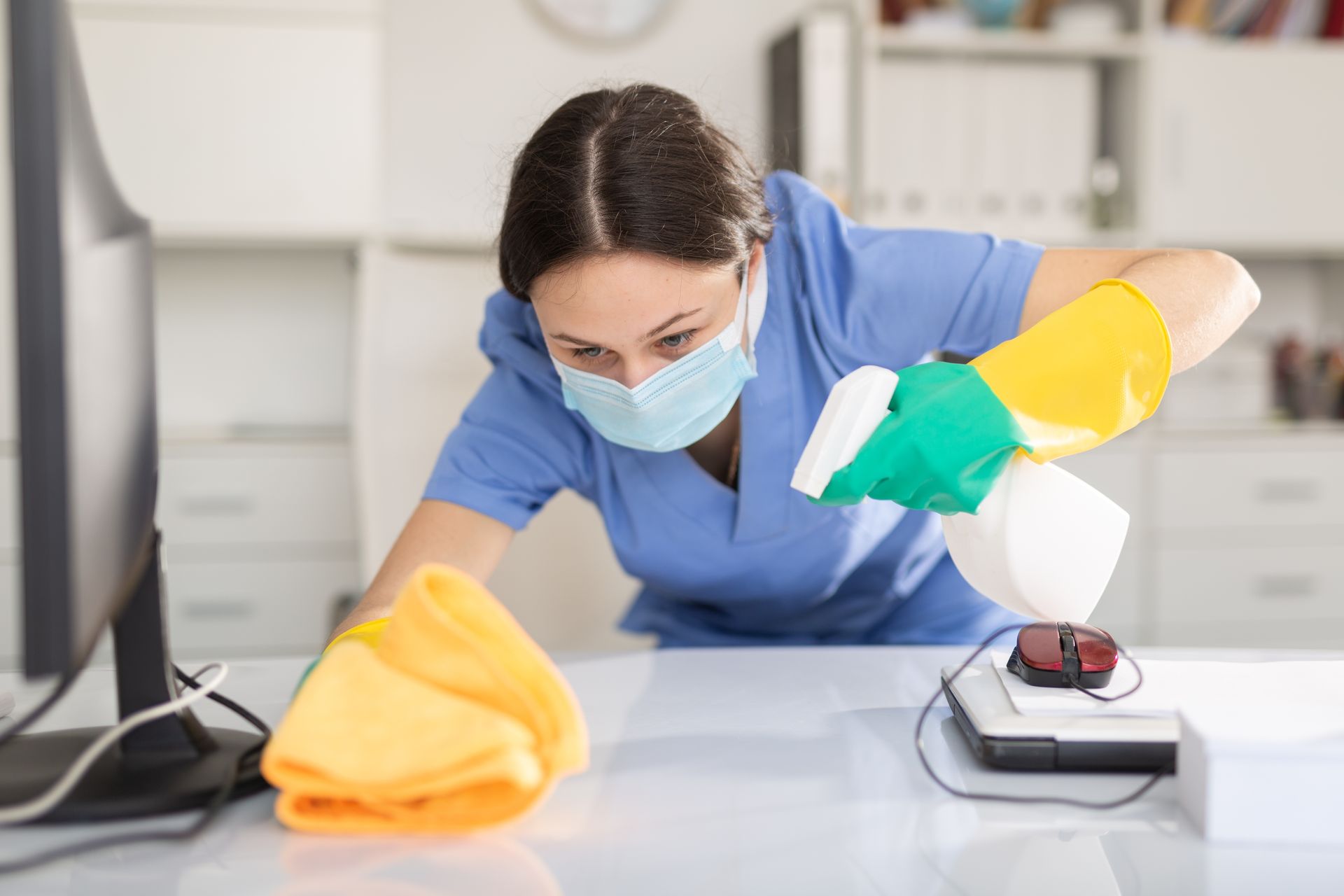 Agent de nettoyage en blouse bleue et masque désinfectant un bureau avec des gants et un vaporisateur dans un environnement de bureau.