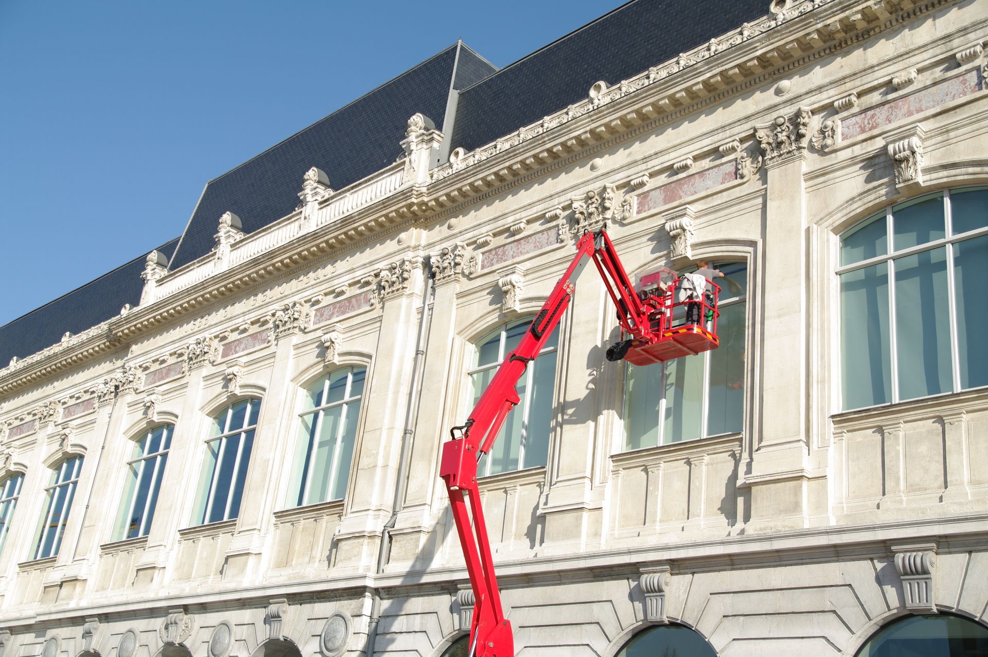 Une nacelle élévatrice rouge est installée à côté de la façade d'un bâtiment historique aux hautes fenêtres et aux ornements en pierre.