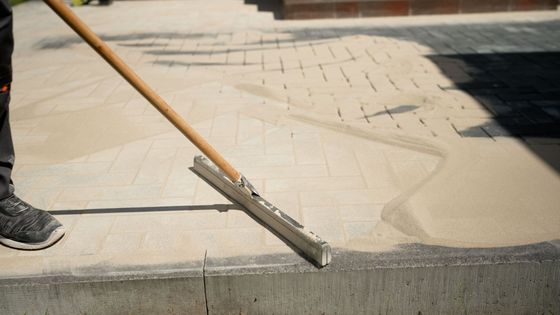 Une personne utilise une brosse à long manche pour nettoyer un trottoir poussiéreux au soleil.