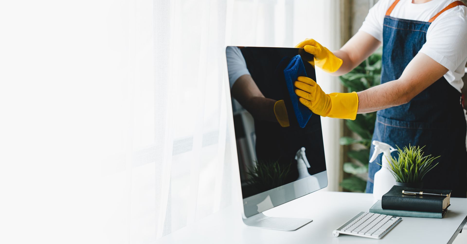 Une personne portant des gants jaunes nettoie un écran d'ordinateur à un bureau où se trouvent un clavier et une plante.