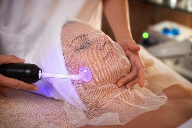 Mujer recibe un tratamiento facial de alta frecuencia en un spa, con un dispositivo que emite una luz violeta y una red de malla sobre su cara.