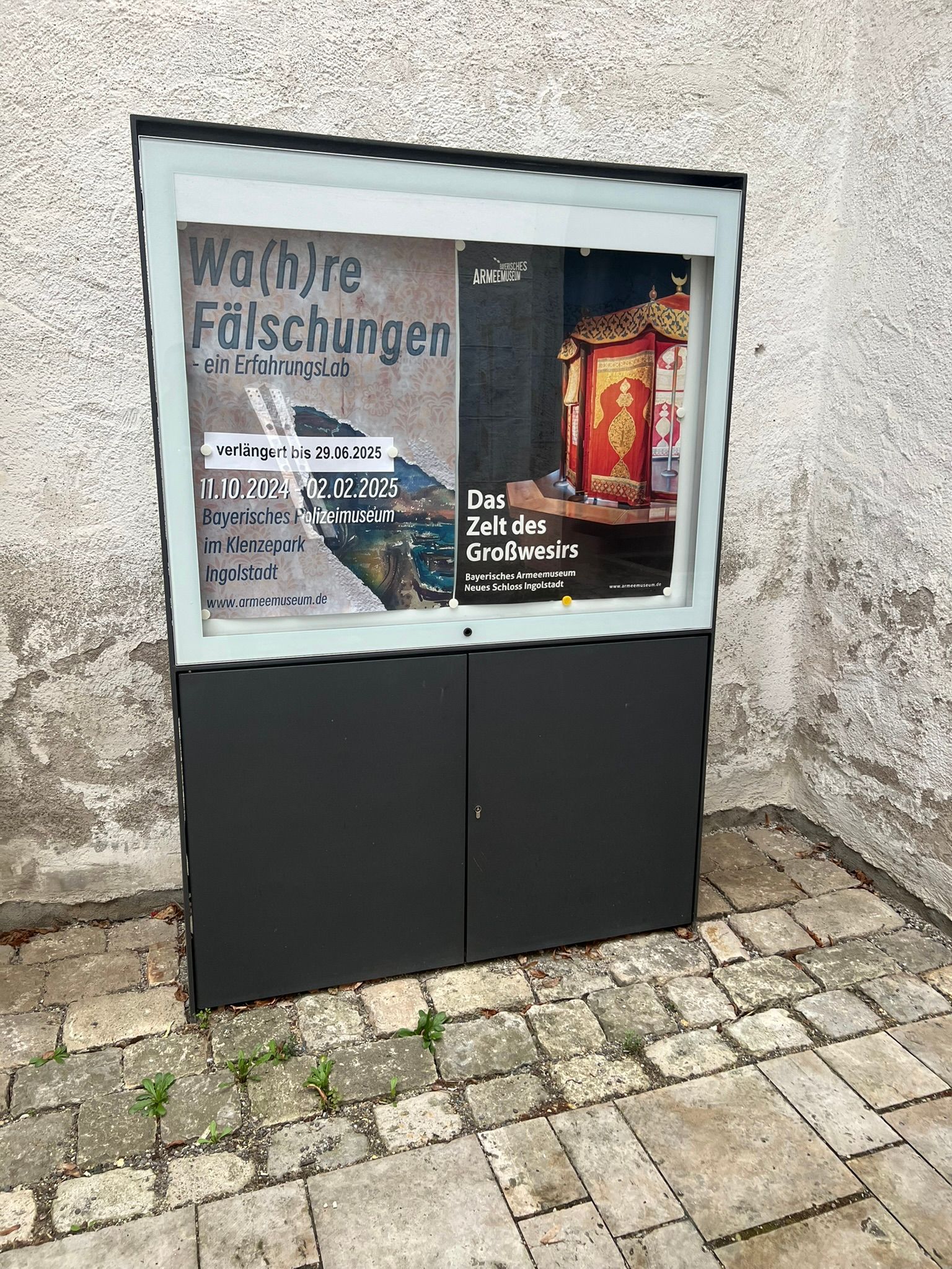 Auf einem gepflasterten Gehweg steht eine Vitrine mit einem Poster darauf.