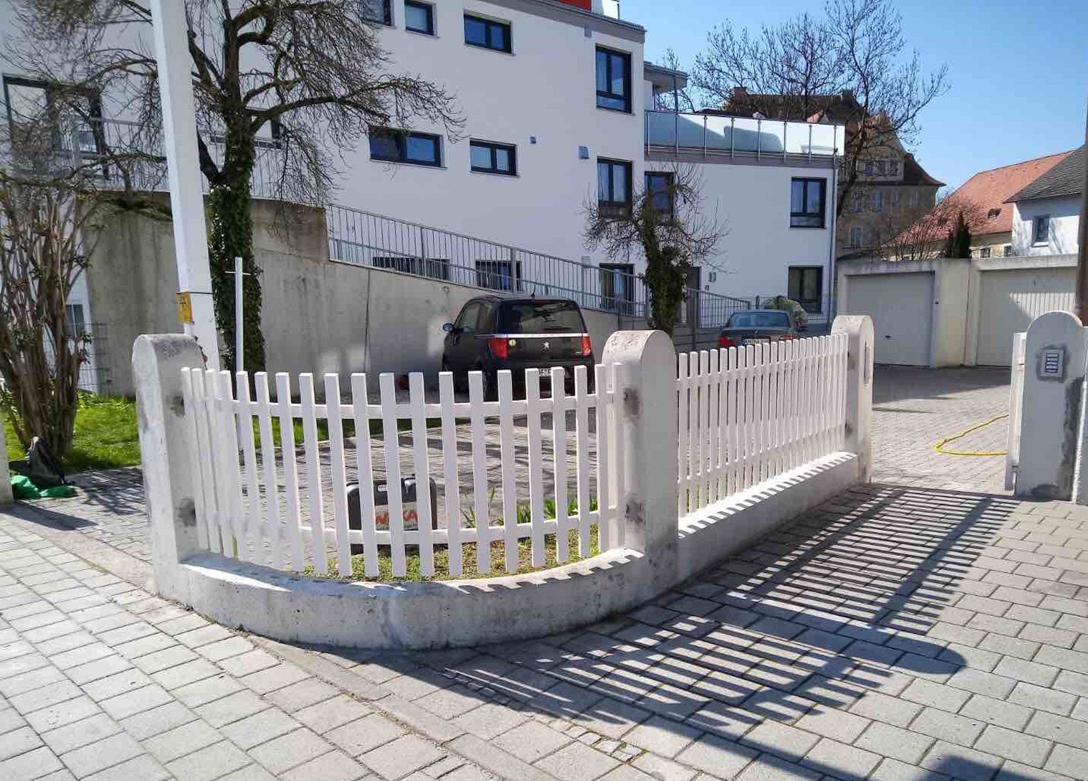Ein weißer Lattenzaun umgibt einen Parkplatz vor einem Gebäude.