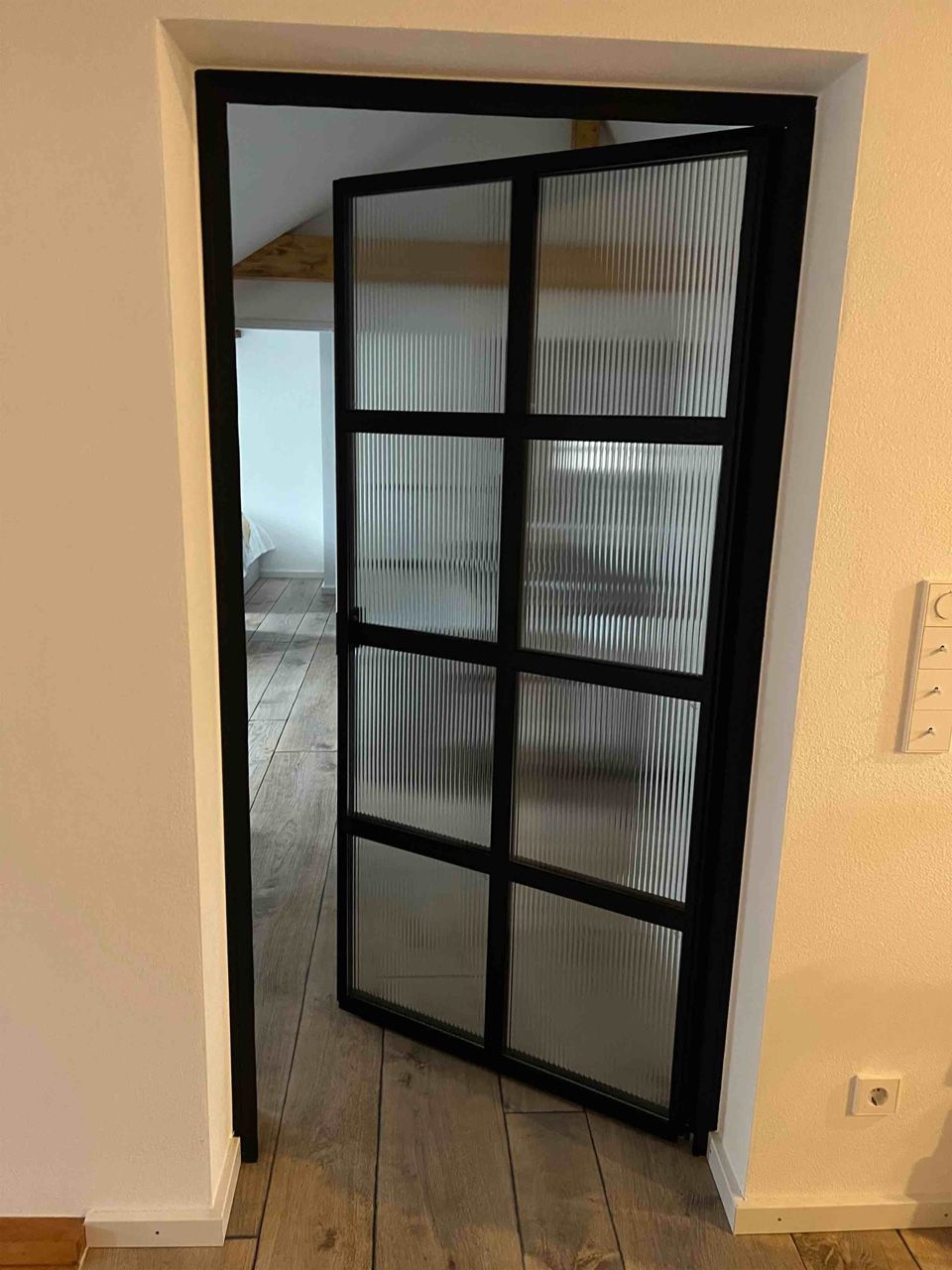 In einem Raum steht eine Glastür mit schwarzem Rahmen offen.