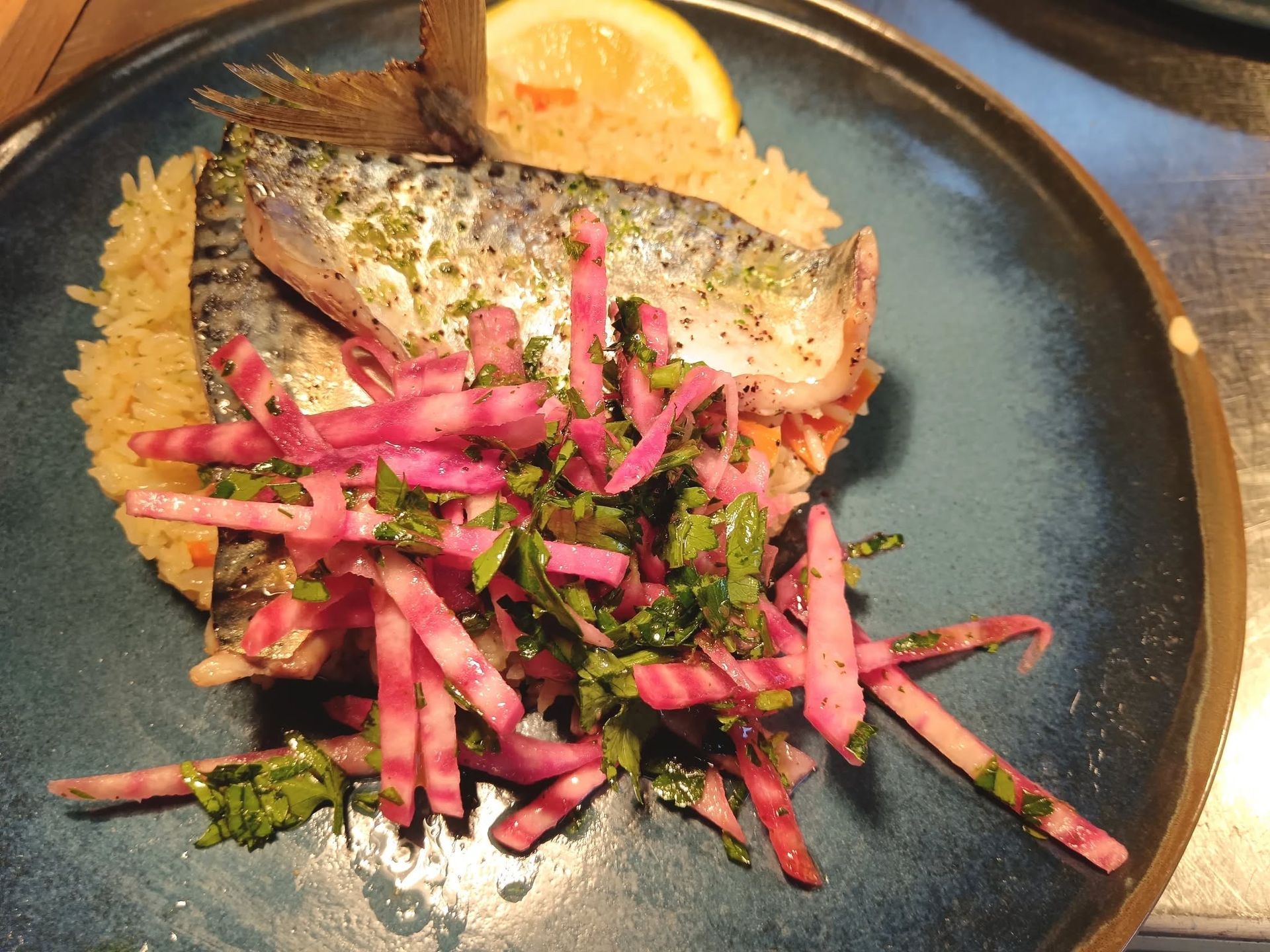Poisson grillé avec du riz, une tranche de citron et des légumes marinés présenté dans une assiette bleue foncée.