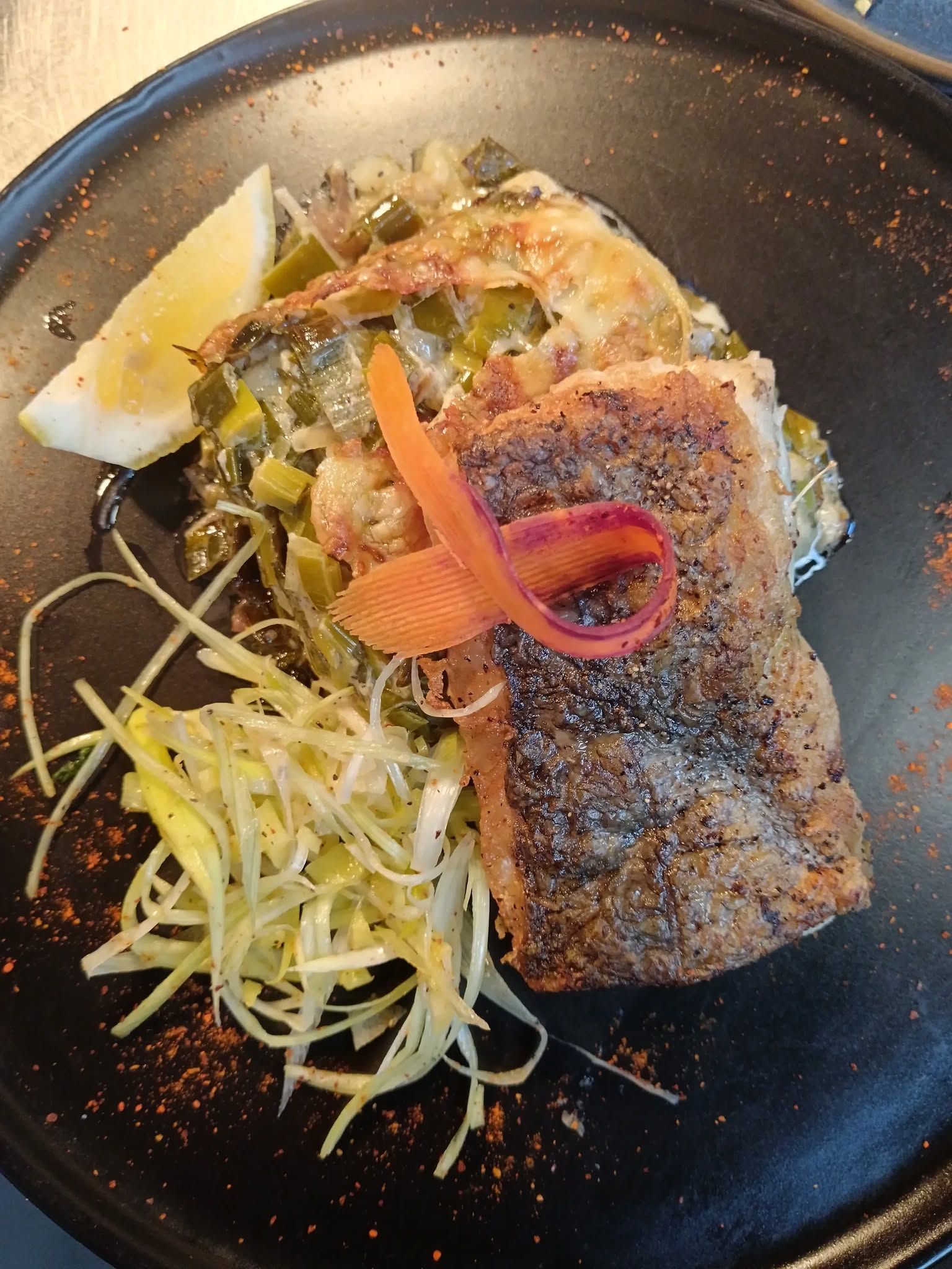 Poisson poêlé accompagné de légumes râpés, d'oignons marinés et d'un quartier de citron présenté dans une assiette noire.
