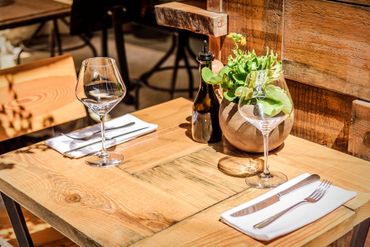 Table de restaurant en bois dressée avec des verres à vin, des couverts, des serviettes et un centre de table composé d'un pot d'herbes aromatiques.