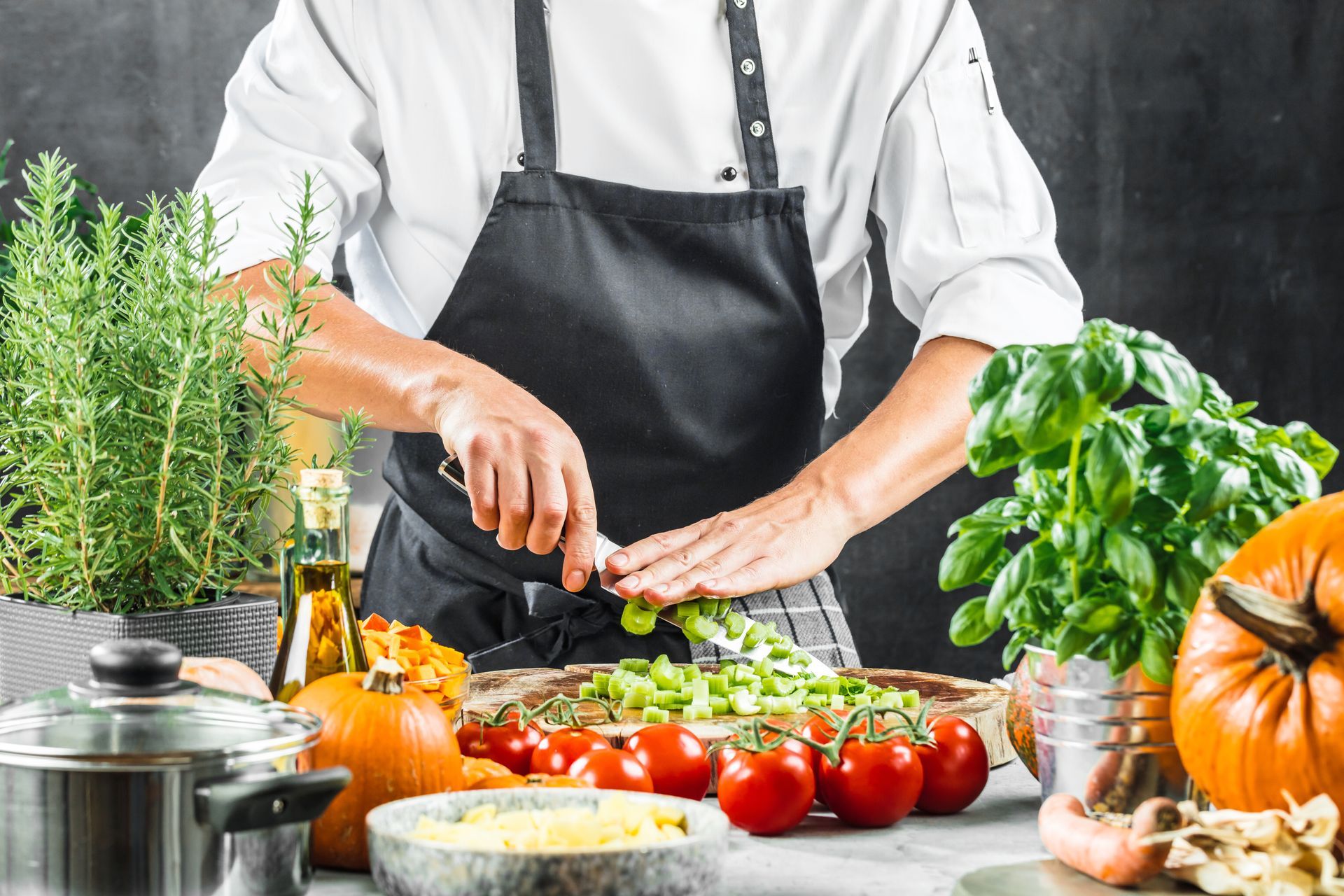 Un chef émince des herbes aromatiques, entouré de tomates, de basilic, de courges et de divers ustensiles de cuisine.
