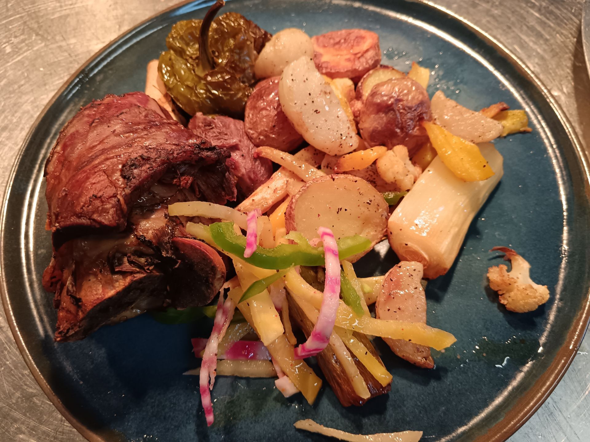Assiette composée de viande grillée, de légumes rôtis, de pommes de terre et d'oignons marinés, servie dans un plat bleu.