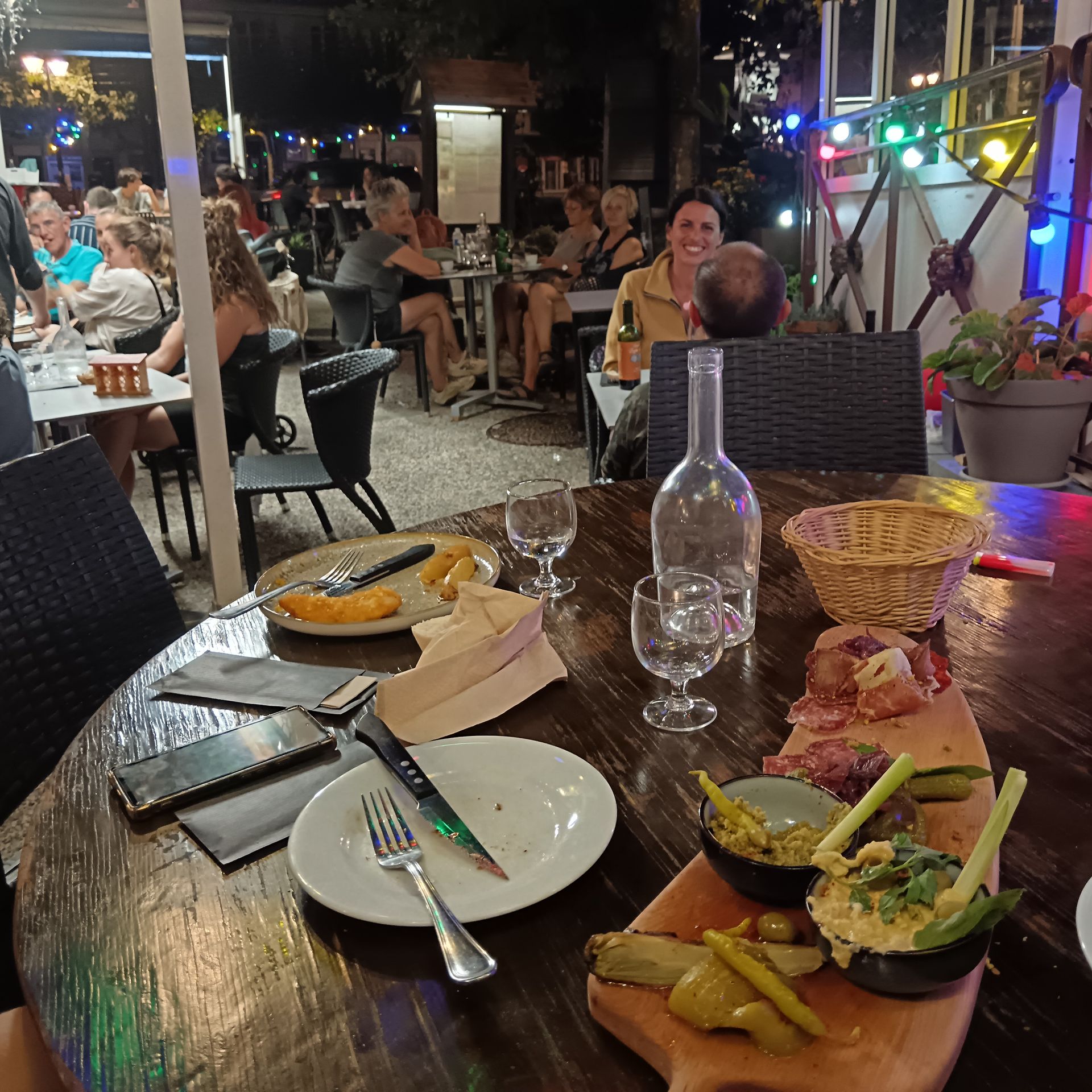Table de restaurant en terrasse avec assiettes et verres vides, donnant sur une salle à manger animée le soir.