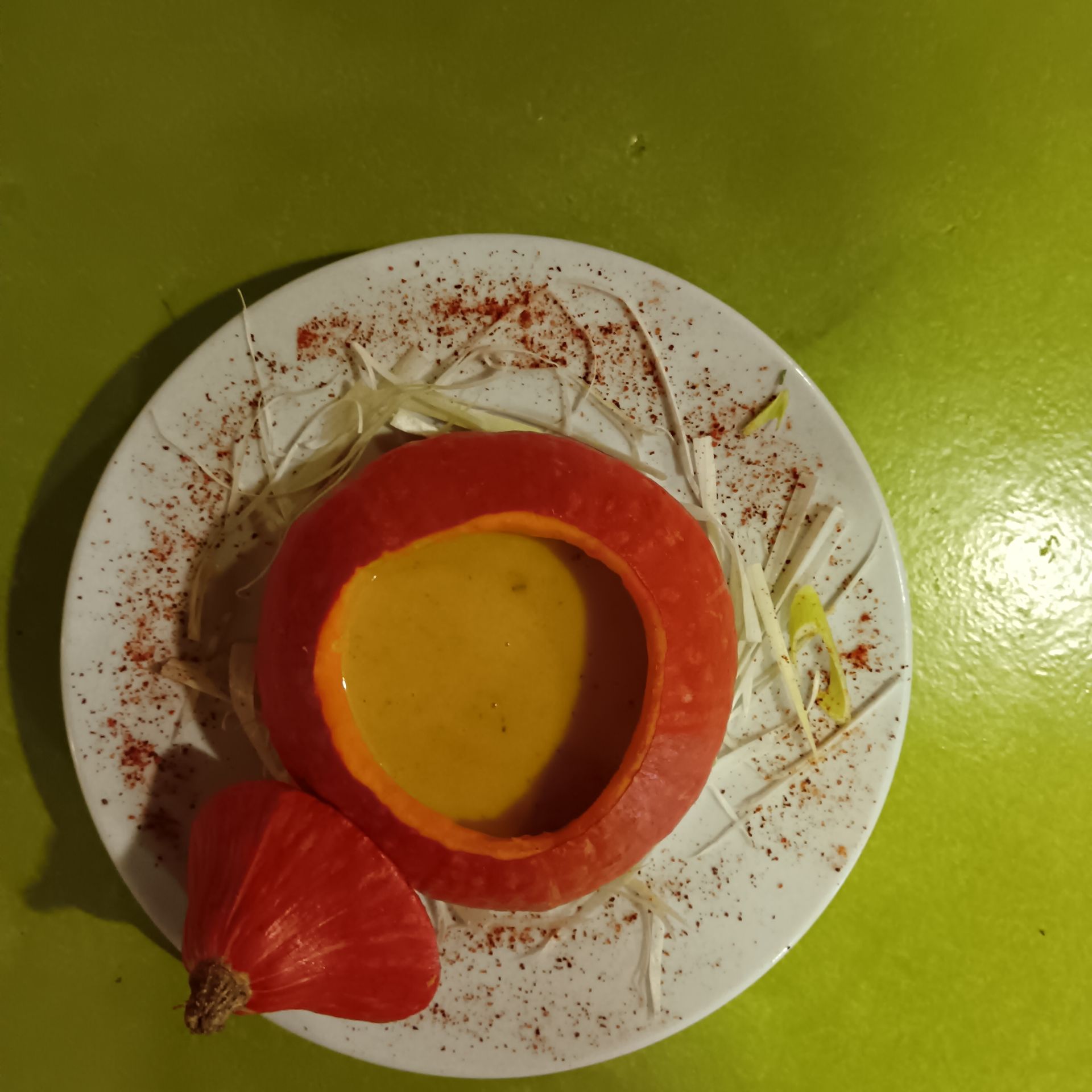 Soupe de potiron servie dans une citrouille évidée sur une assiette blanche, garnie de fromage râpé et d'épices, avec un quartier de potiron.