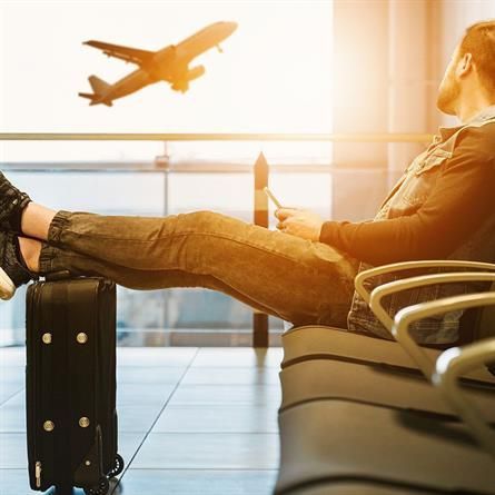 Ein Mann sitzt im Wartebereich eines Flughafens, seine Füße stehen auf einem Koffer, im Hintergrund fliegt ein Flugzeug.