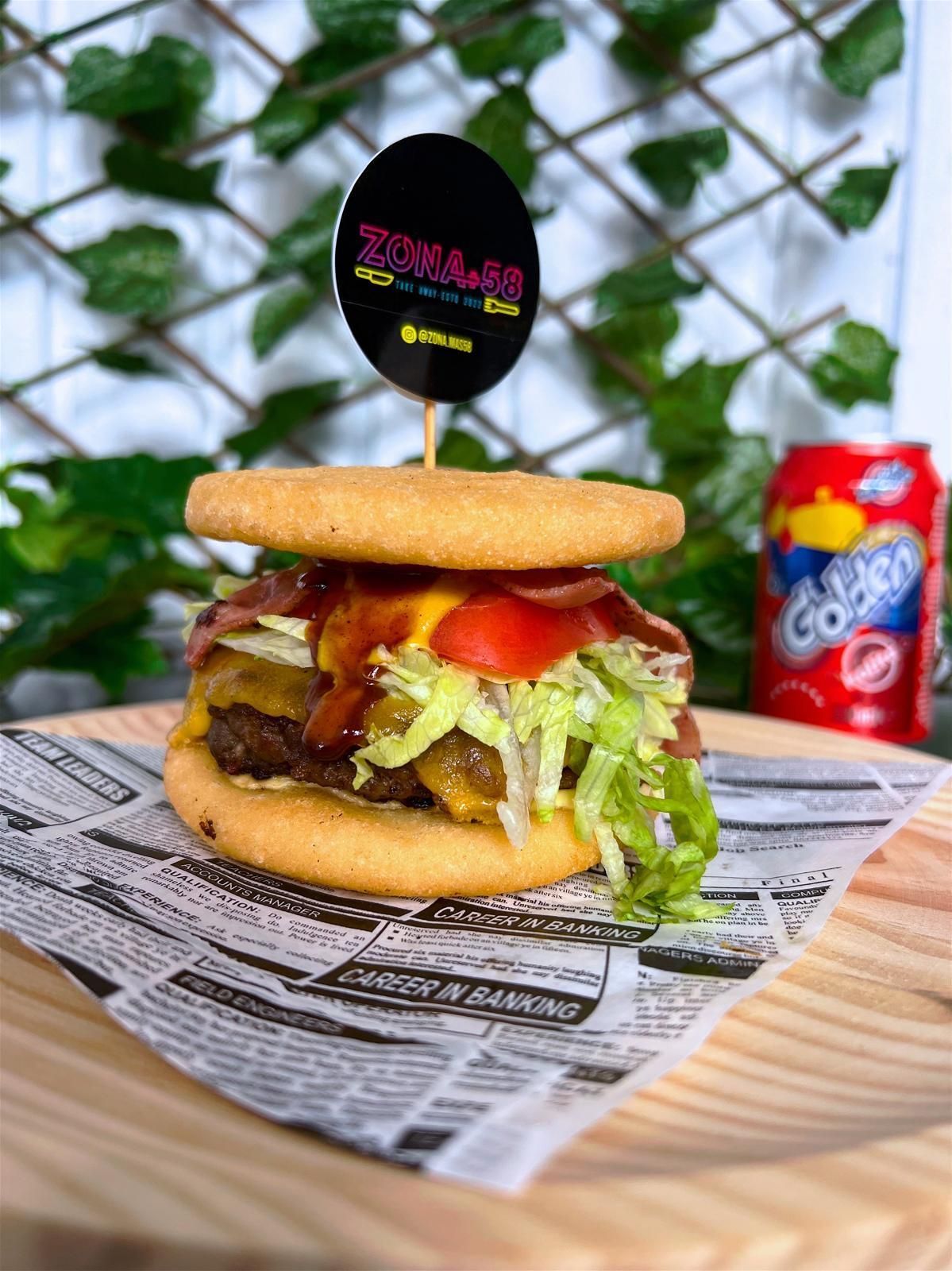 Una hamburguesa está sobre una tabla de cortar de madera al lado de una lata de refresco.