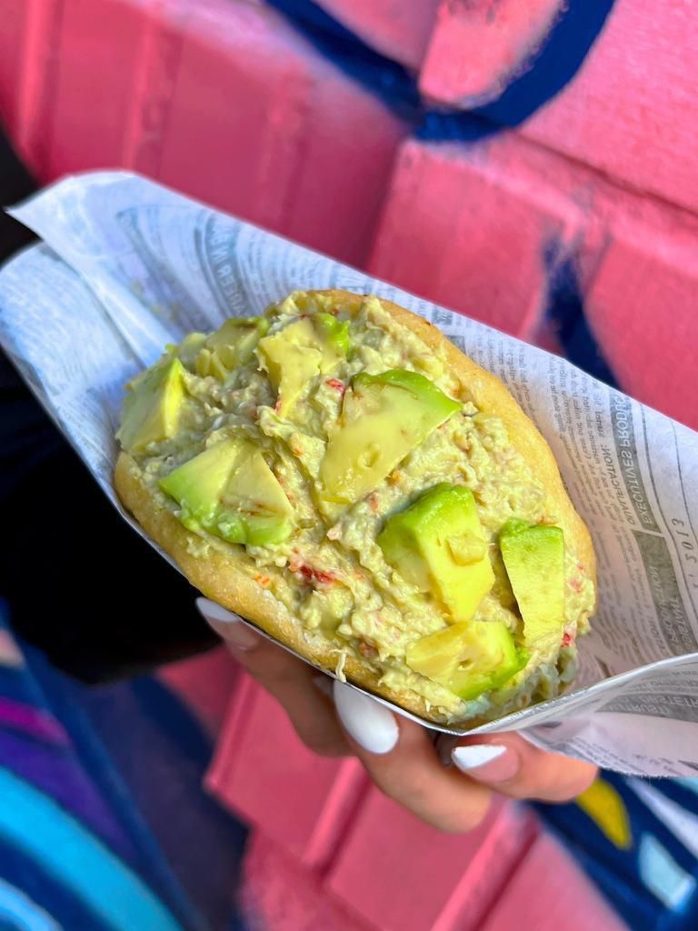 Una persona sostiene un sándwich con aguacate delante de una pared rosa.