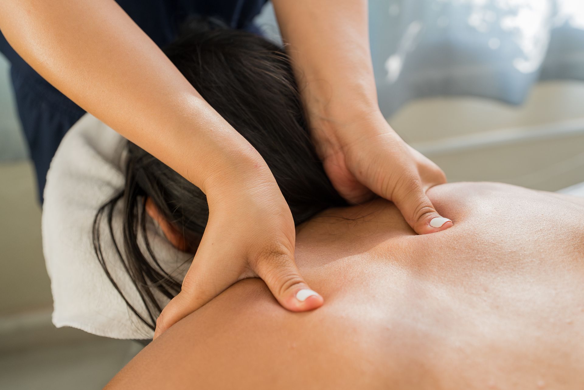 Hände drücken während einer Massage auf den nackten Rücken einer Person, im Hintergrund eine verschwommene Spa-Kulisse.