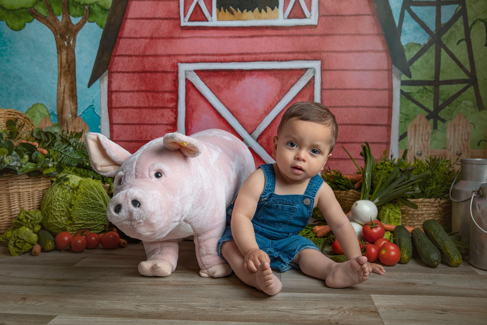 Un bebé está sentado junto a un cerdo de peluche frente a un granero.