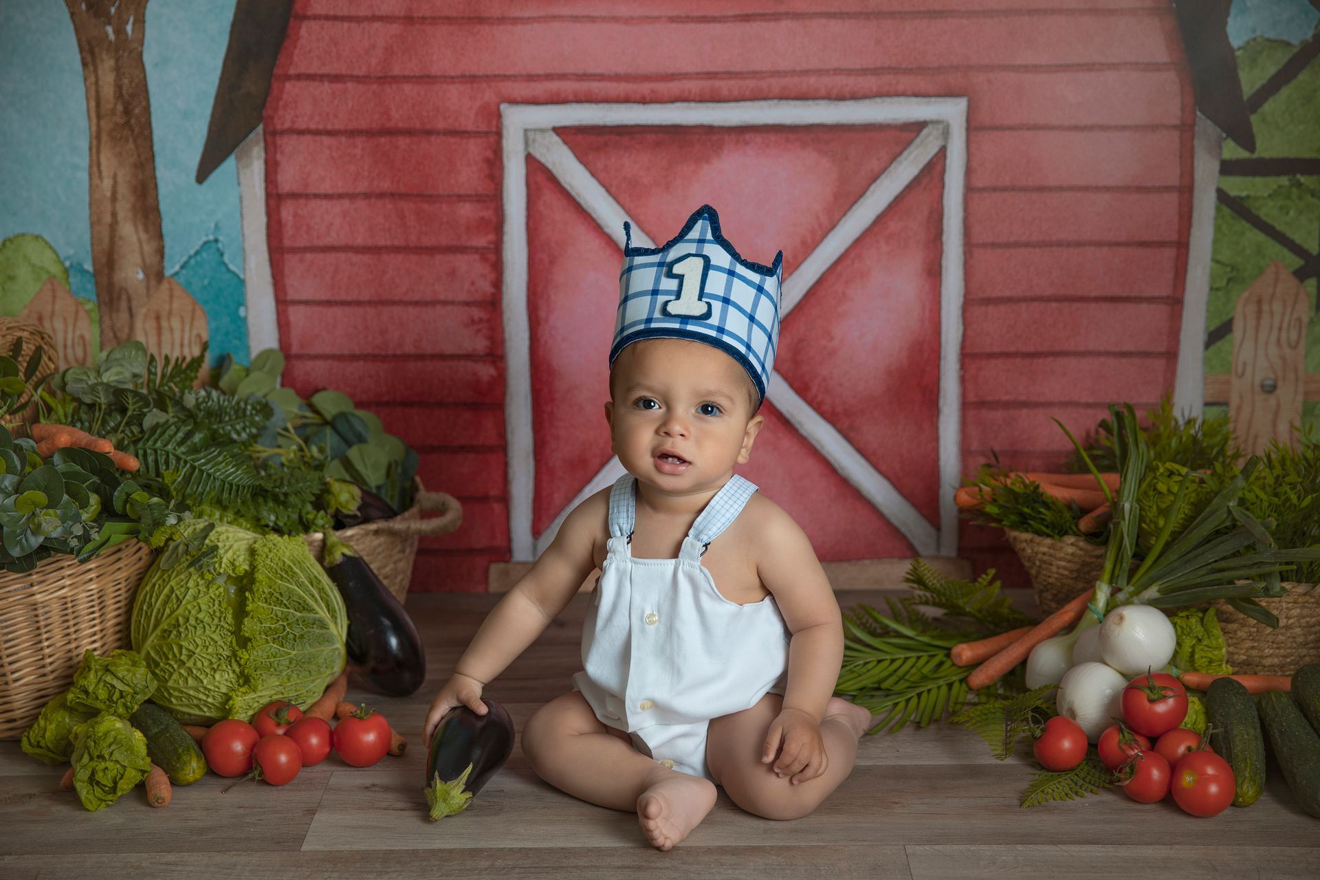Un bebé que lleva una corona está sentado frente a un granero rodeado de verduras.