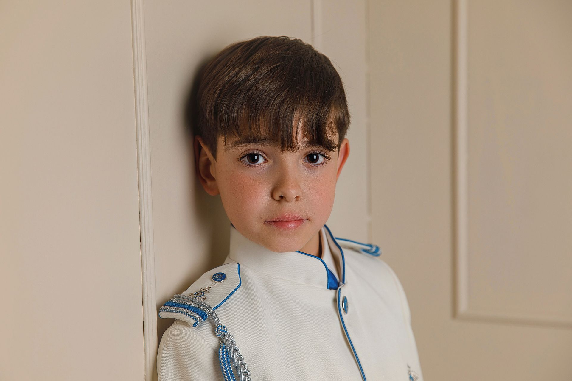 Un niño pequeño con un traje blanco está apoyado contra una pared.
