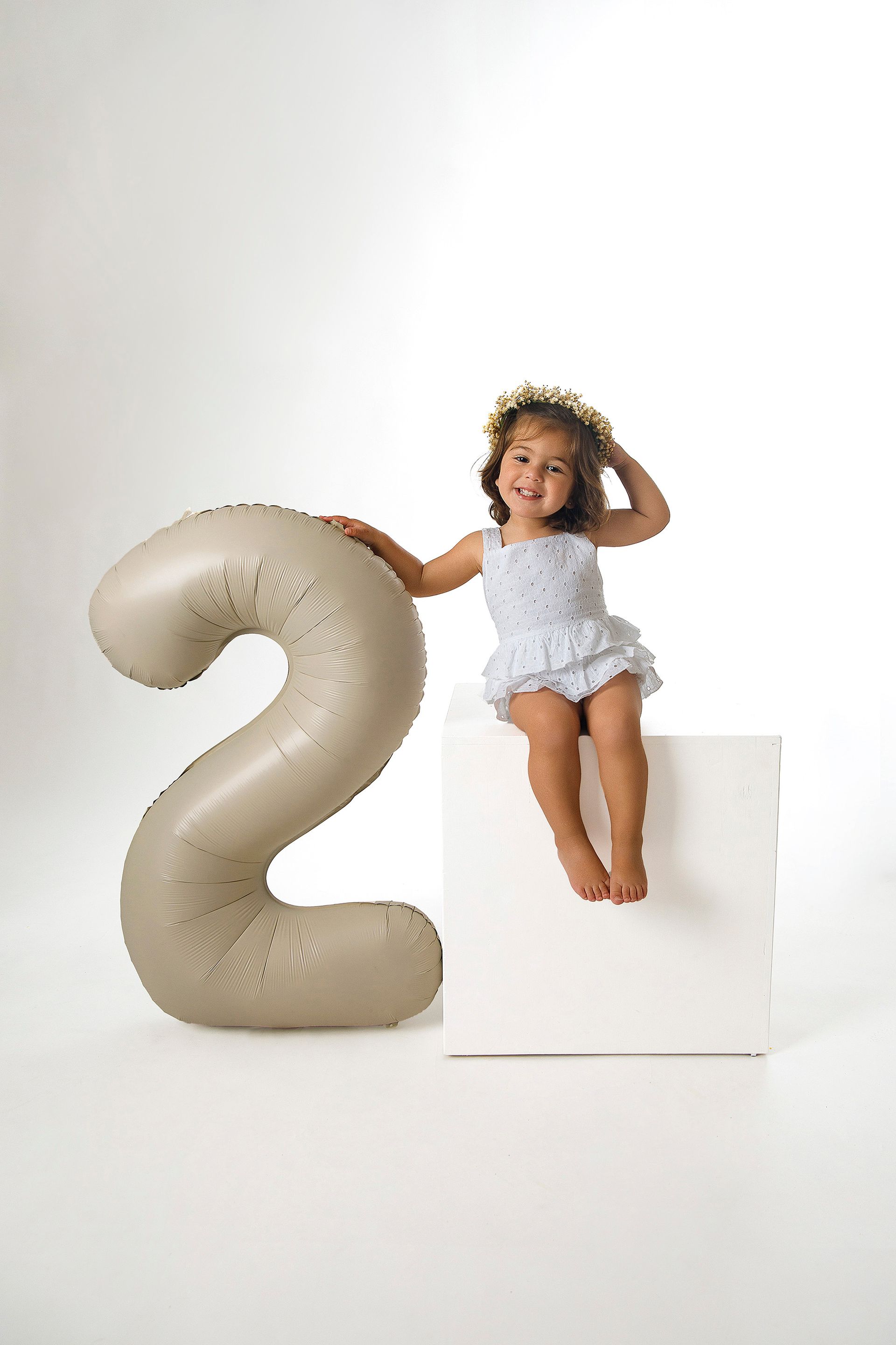 Una niña está sentada en una caja al lado de un globo grande con el número dos.