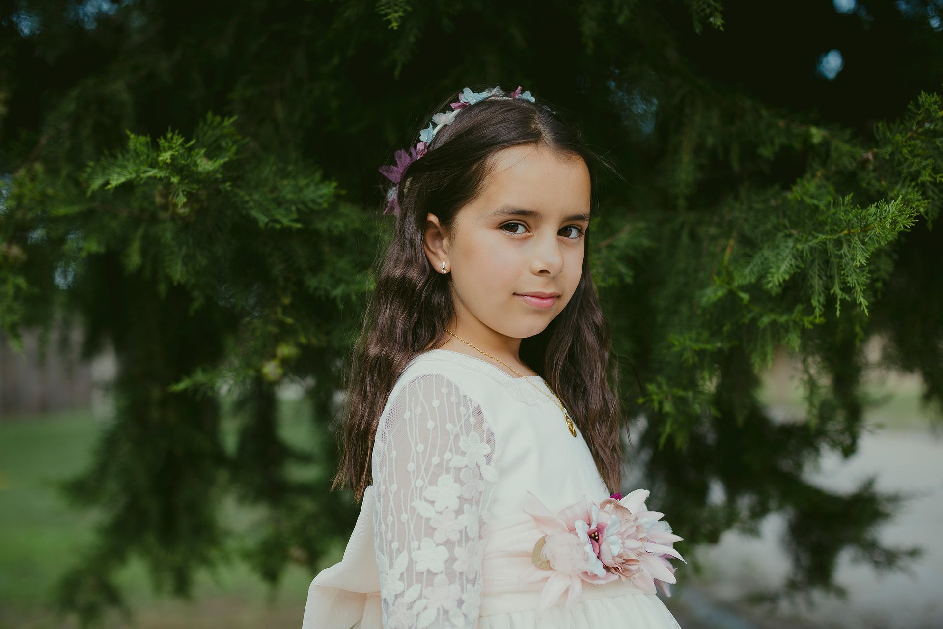 Una niña con un vestido blanco está parada frente a un árbol.