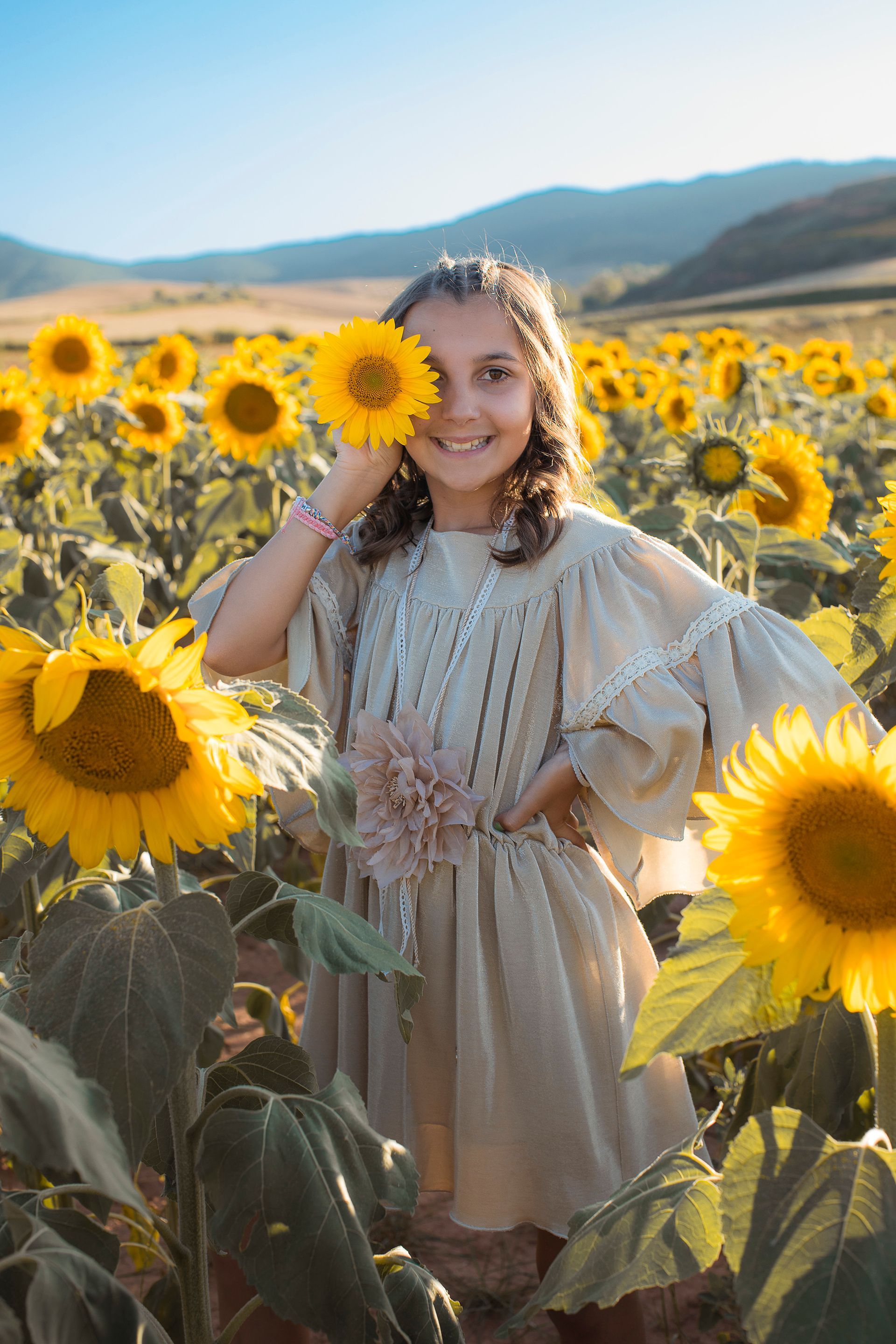 Una niña está parada en un campo de girasoles cubriéndose la cara con un girasol.
