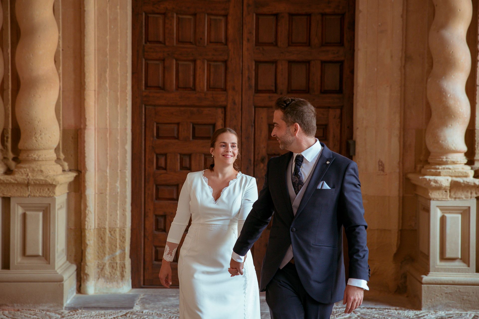 Una novia y un novio caminan frente a una puerta de madera tomados de la mano.