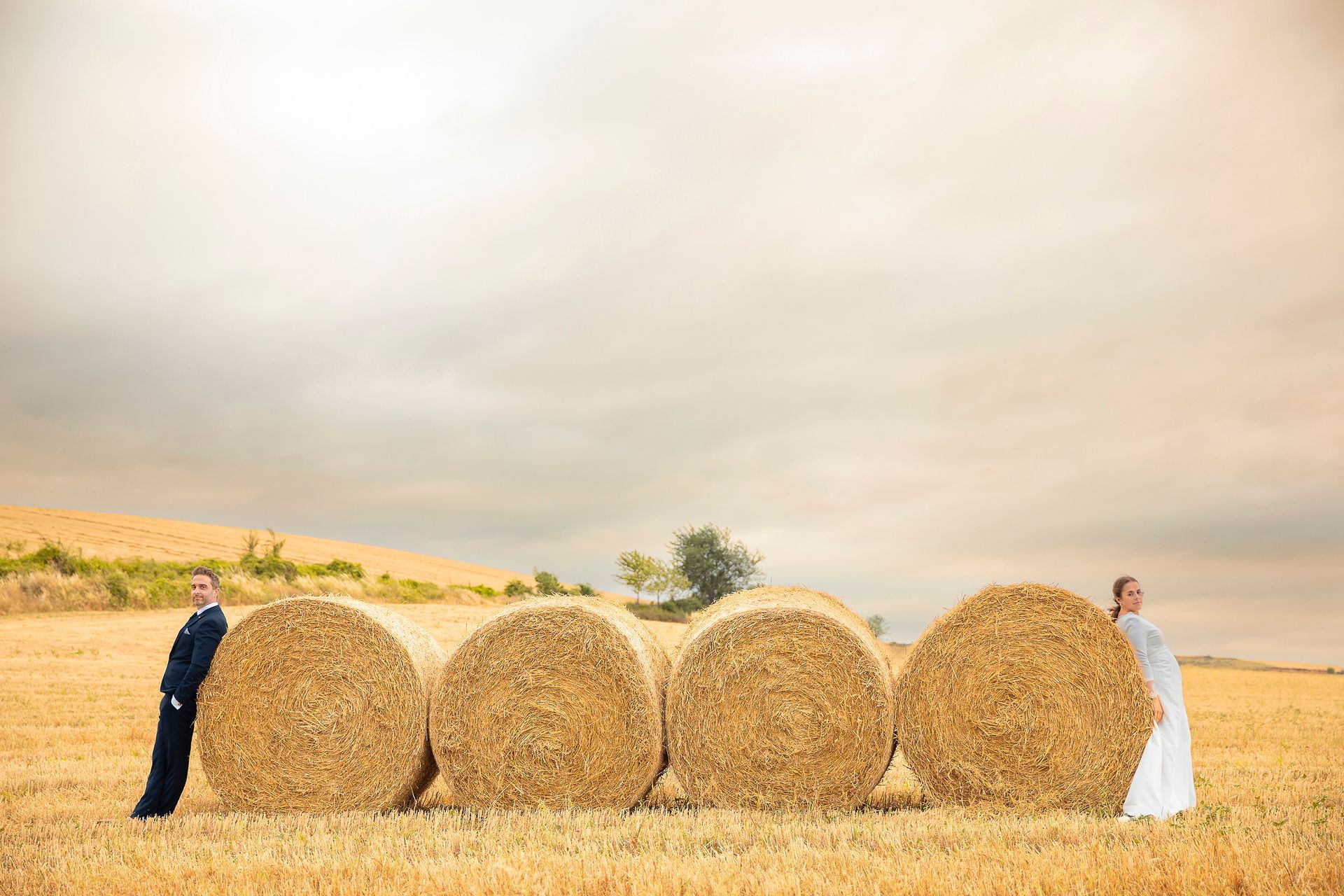 Una novia y un novio están de pie junto a fardos de heno en un campo.