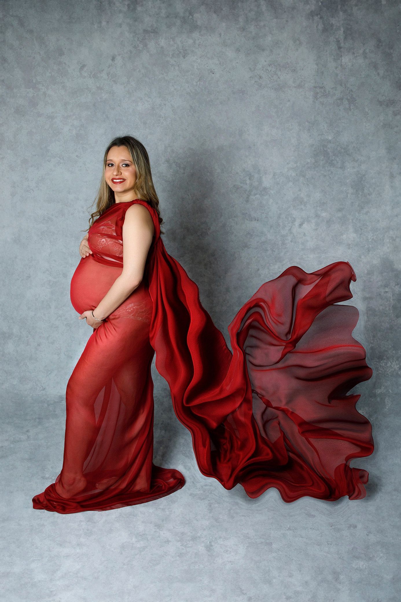 Una mujer embarazada con un vestido rojo está parada frente a un fondo gris.